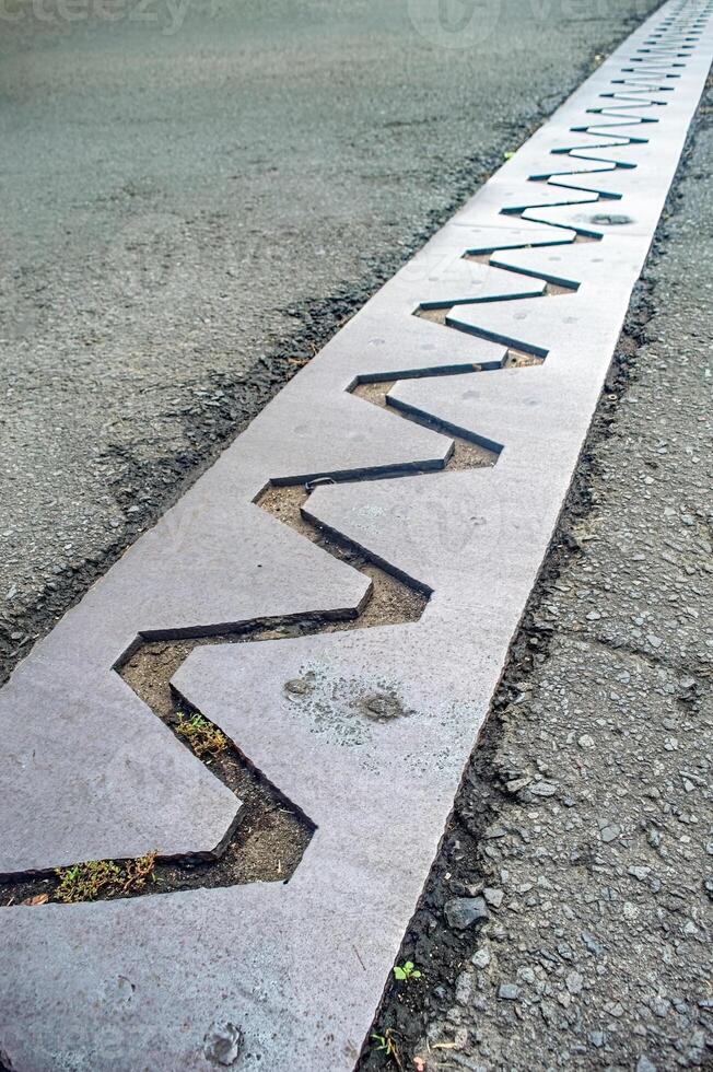 A close up view of a bridge expansion joint with a zigzag pattern, designed to accommodate structural movements and prevent damage. The metal plate is embedded in the asphalt road surface photo