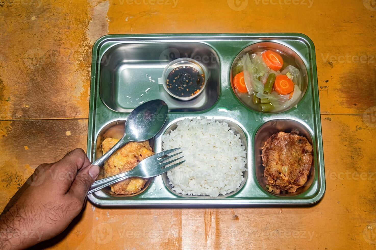 A stainless steel lunch tray with a nutritious meal consisting of rice, fried fish, an omelette, and a small container of sauce. A hand is holding a spoon and fork, ready to eat photo