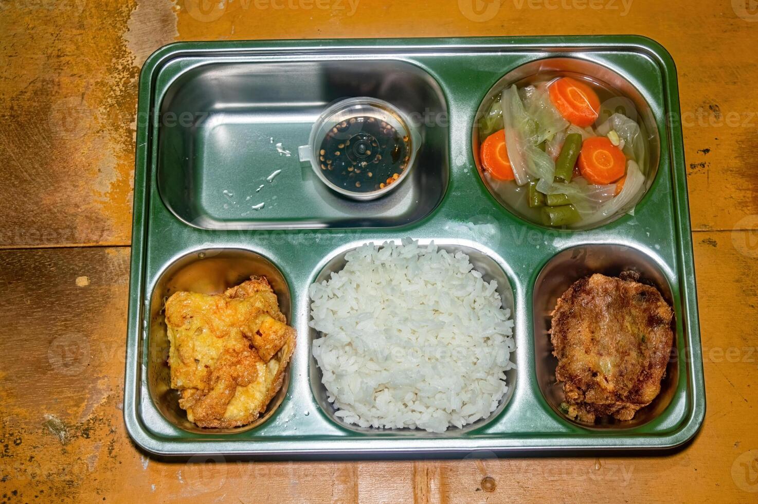 A nutritious and balanced free lunch featuring rice, fried chicken, vegetable soup, and a dipping sauce, served in a stainless steel tray on a rustic wooden table photo
