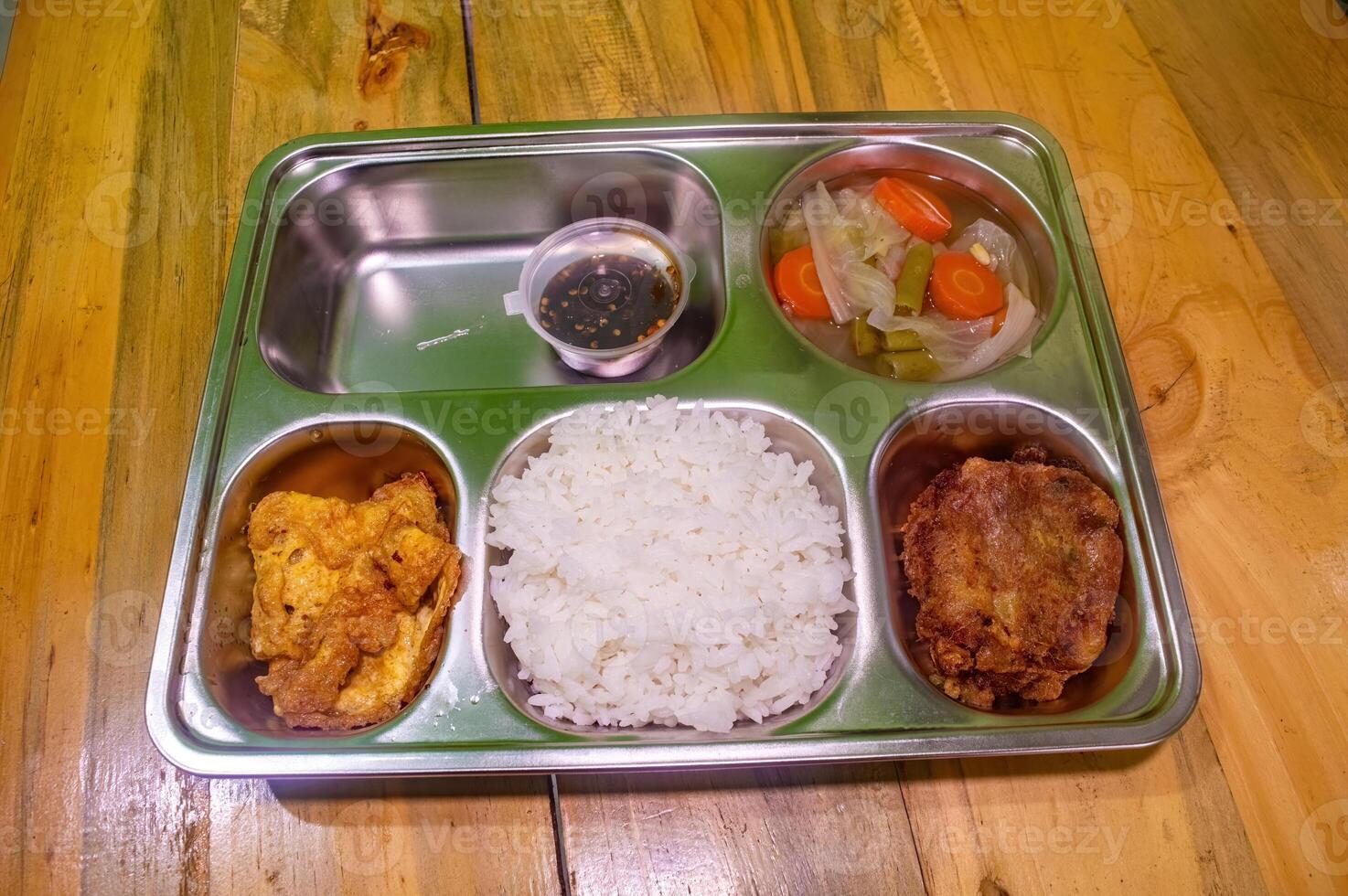 A nutritious free lunch served in a stainless steel tray, featuring rice, fried chicken, an omelette, mixed vegetables, and a small cup of dipping sauce. Perfect for promoting healthy eating photo