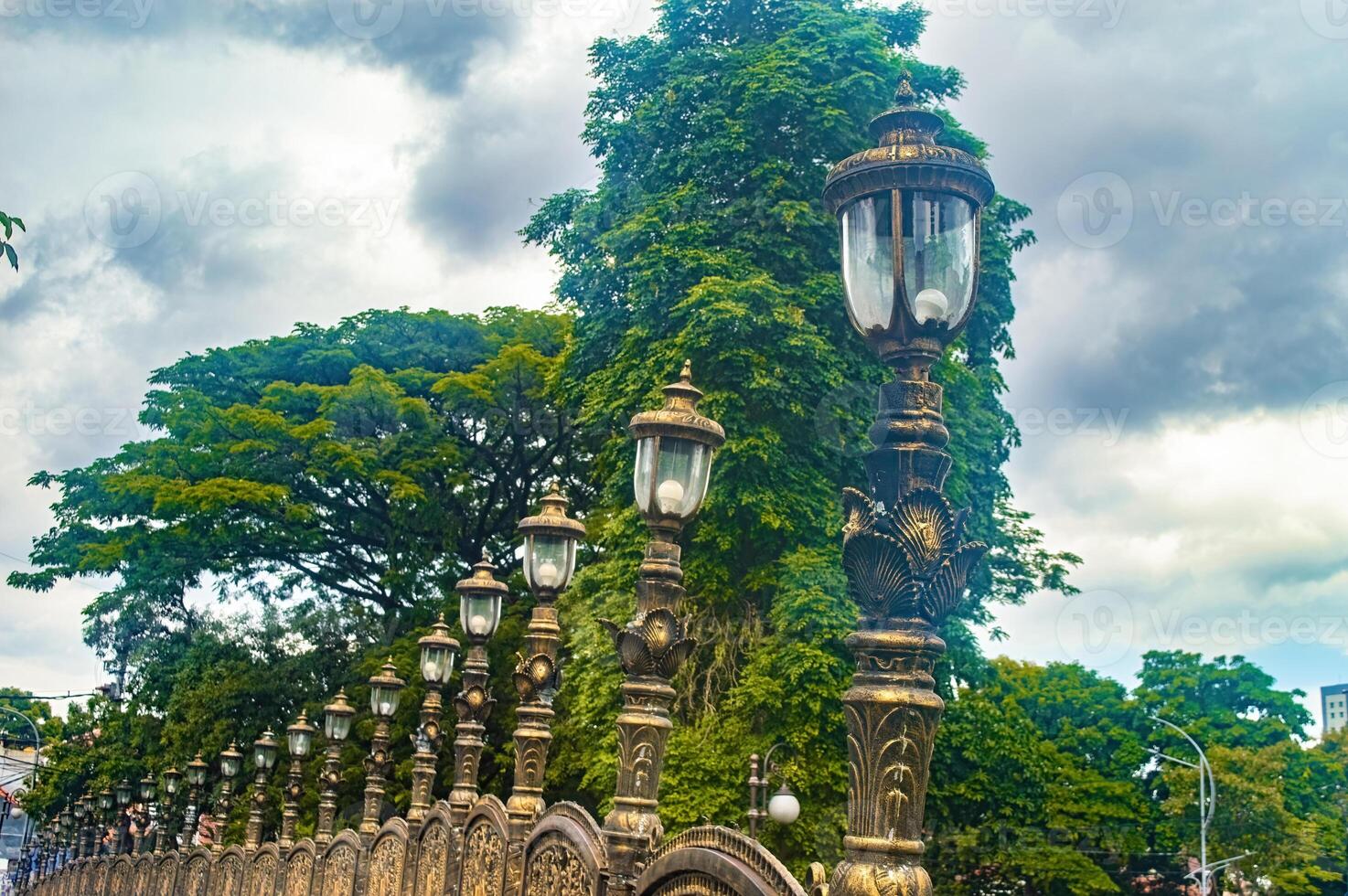 A row of ornate street lamps with intricate designs lines a pathway, set against a backdrop of lush green trees and a cloudy sky, creating a picturesque and historical ambiance photo