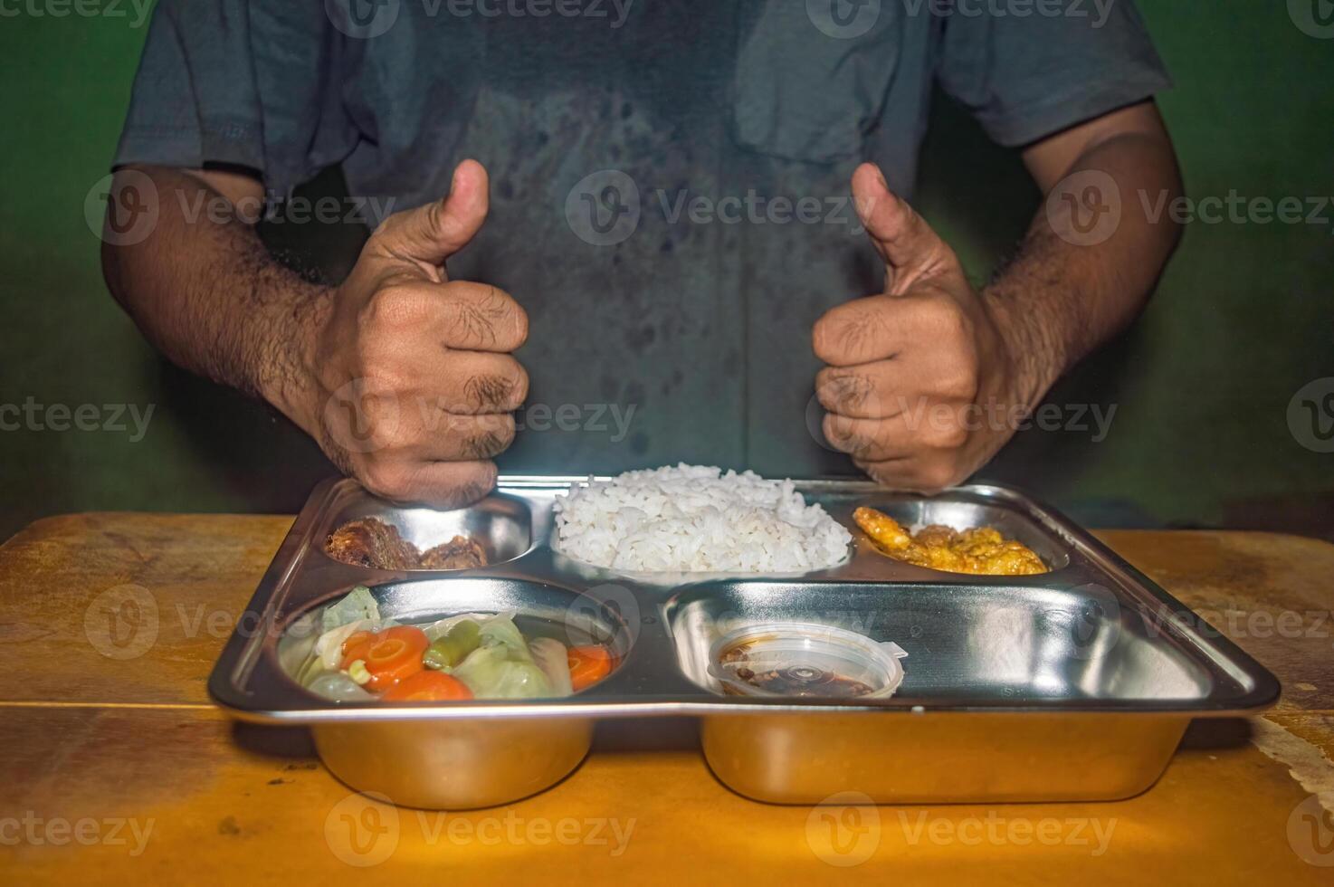 A person gives two thumbs up in front of a stainless steel tray filled with a nutritious meal, including rice, vegetables, and side dishes, symbolizing a healthy and free lunch concept photo