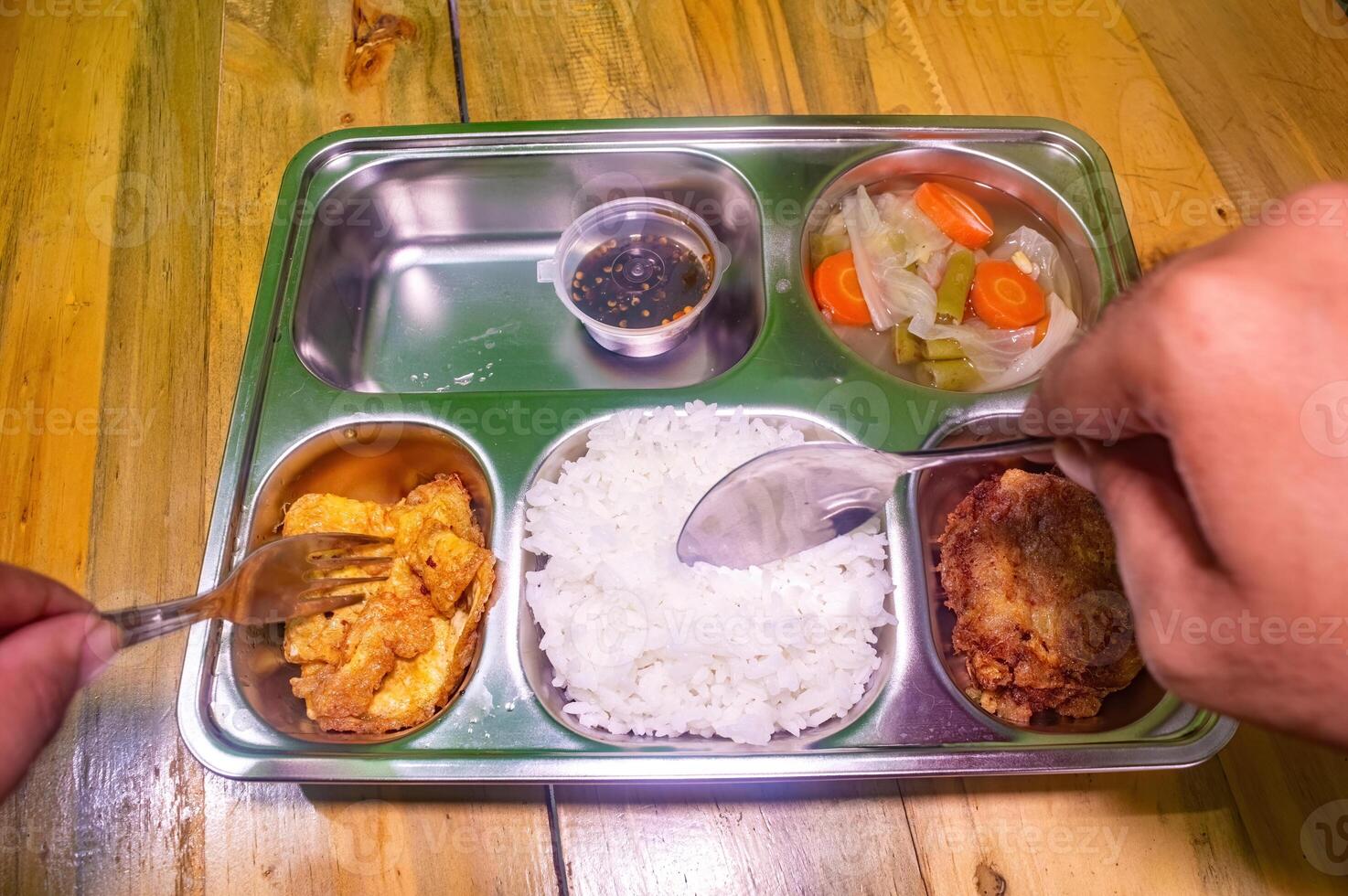A stainless steel lunch tray with rice, fried tofu, vegetables, and a fried patty. Hands are holding a fork and spoon, ready to eat. Ideal for concepts of free or nutritious meals photo