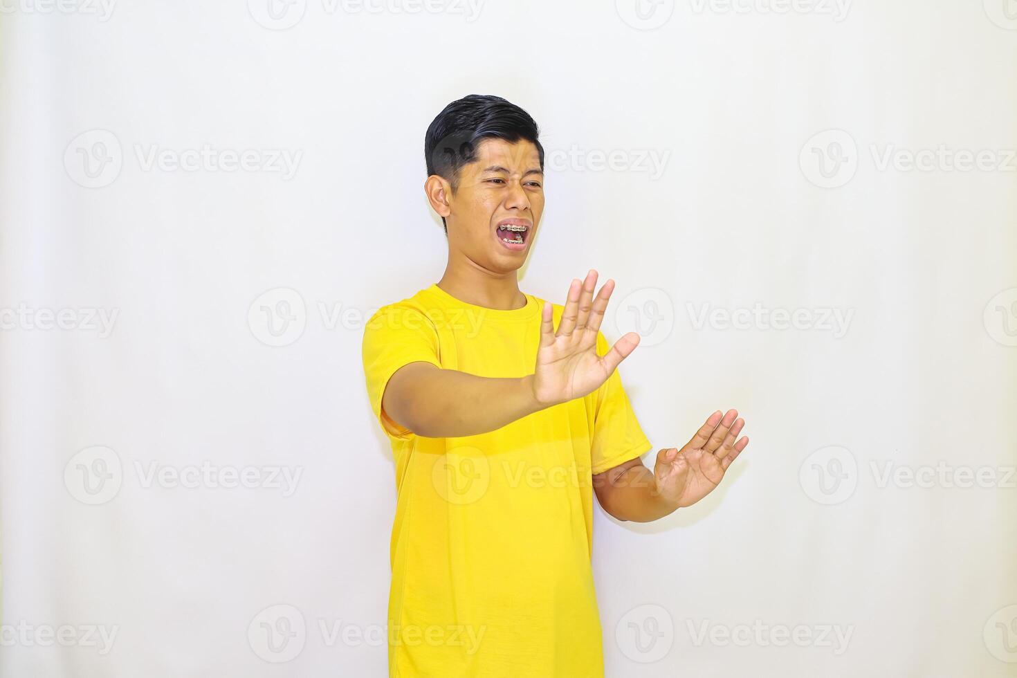 A man in a yellow shirt expresses disgust and rejection with raised hands against a white background, showing an obvious dislike for something, photo