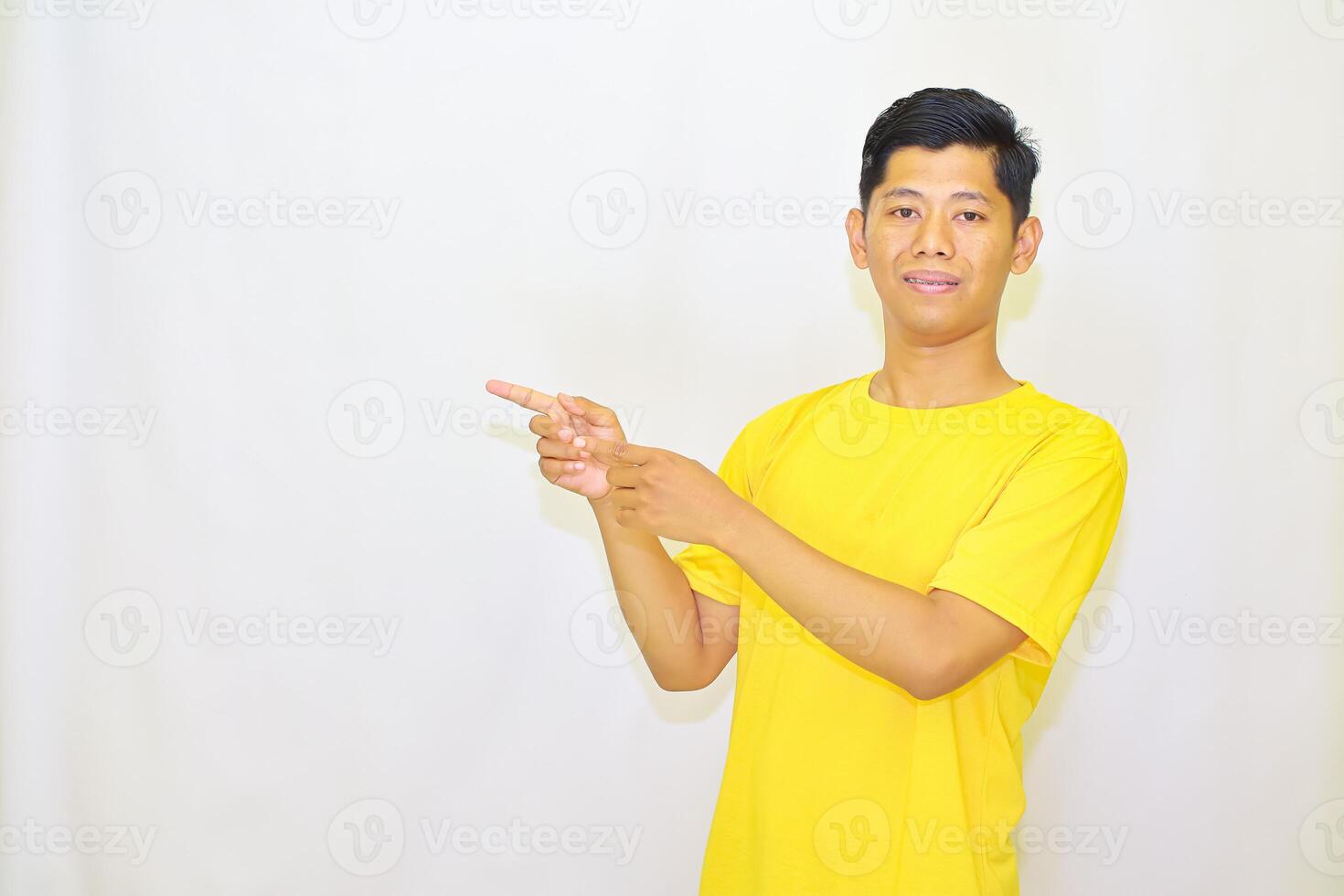 Young man in a yellow t-shirt with a warm smile, pointing towards the left with a focused gaze, showcasing a gesture for announcements or directions, photo