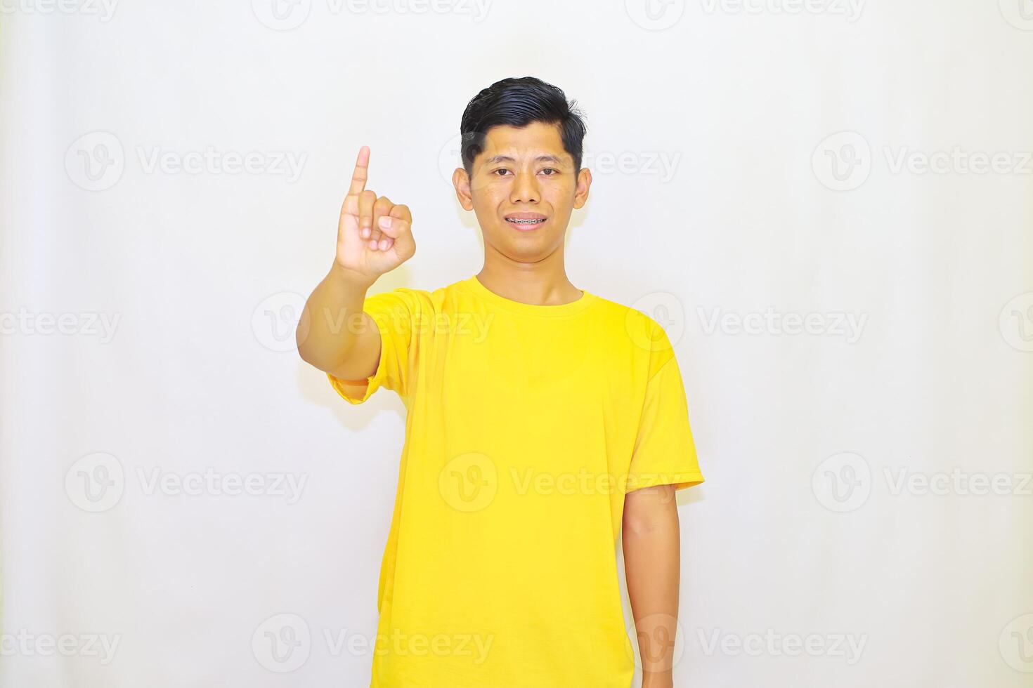 Young man in yellow shirt raising his index finger against a white backdrop, symbolizing importance or the number one, photo