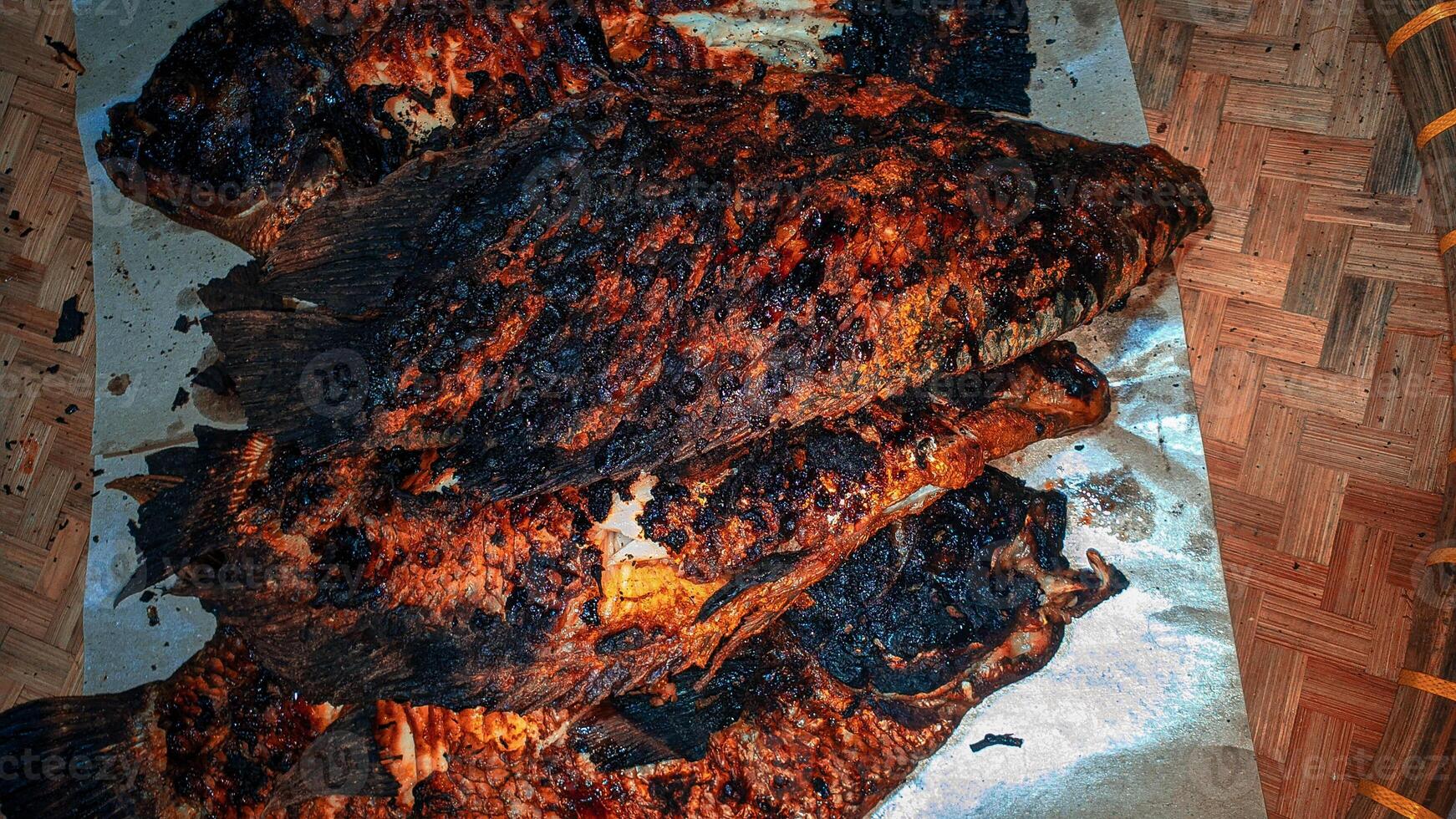 Four whole grilled fish with dark, charred skin, lying on a piece of paper, placed on a woven mat photo