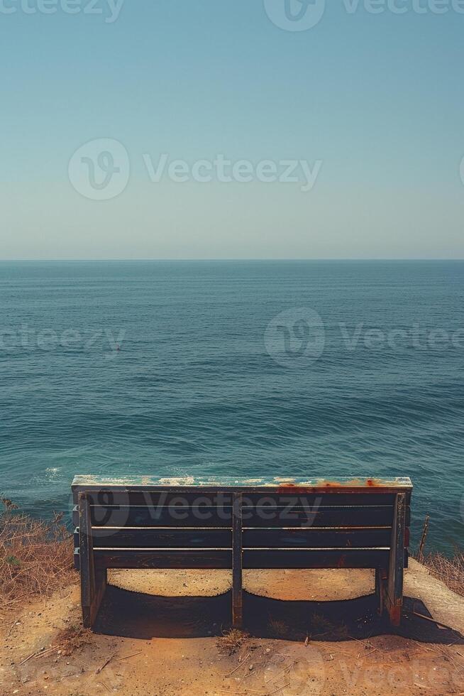 A solitary weathered bench overlooks the vast ocean under a clear blue sky. photo