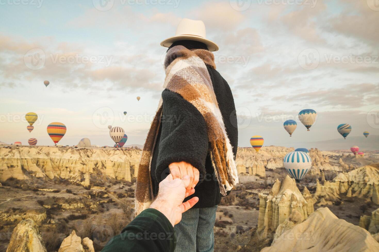 Couple hold hand show looking admire hot air balloon show ,wear fashionable clothing stand on scenic viewpoint watch hot air balloon flight on sunrise with beautiful goreme town and valley background photo