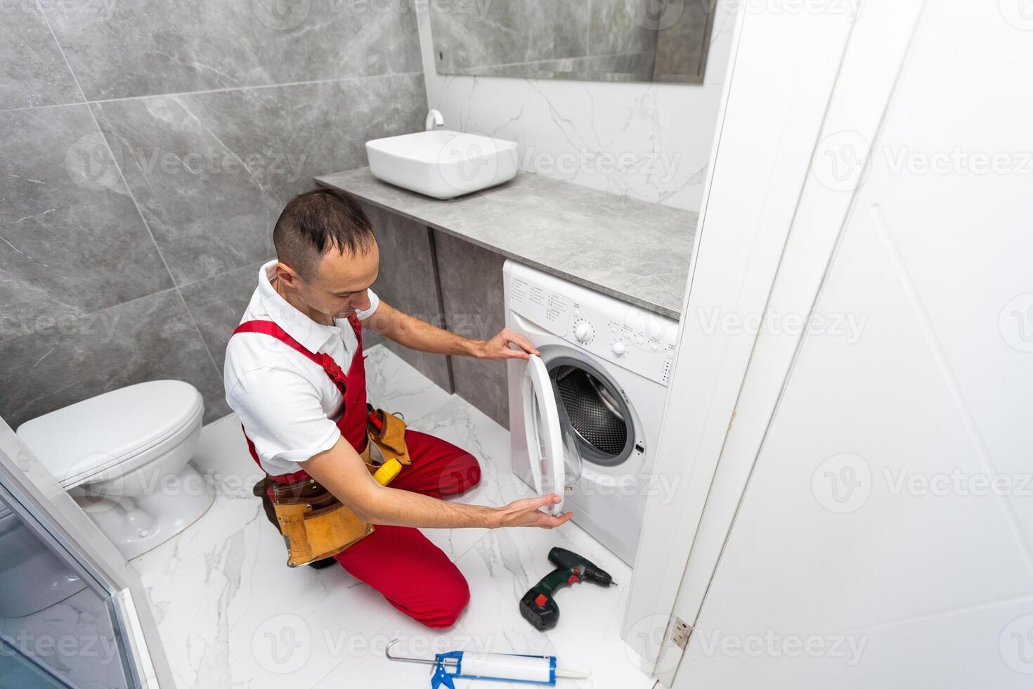 Young smiling happy man plumber handyman in overalls repair fix the washing machine in the kitchen and look to the camera photo