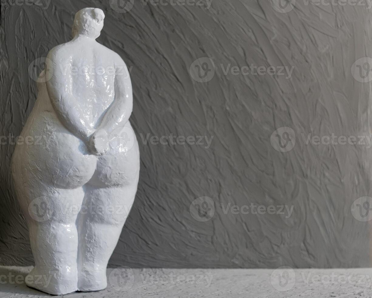 A white plaster figurine of a plump woman turned away from us, set against a gray background with copy space in a minimalist style photo