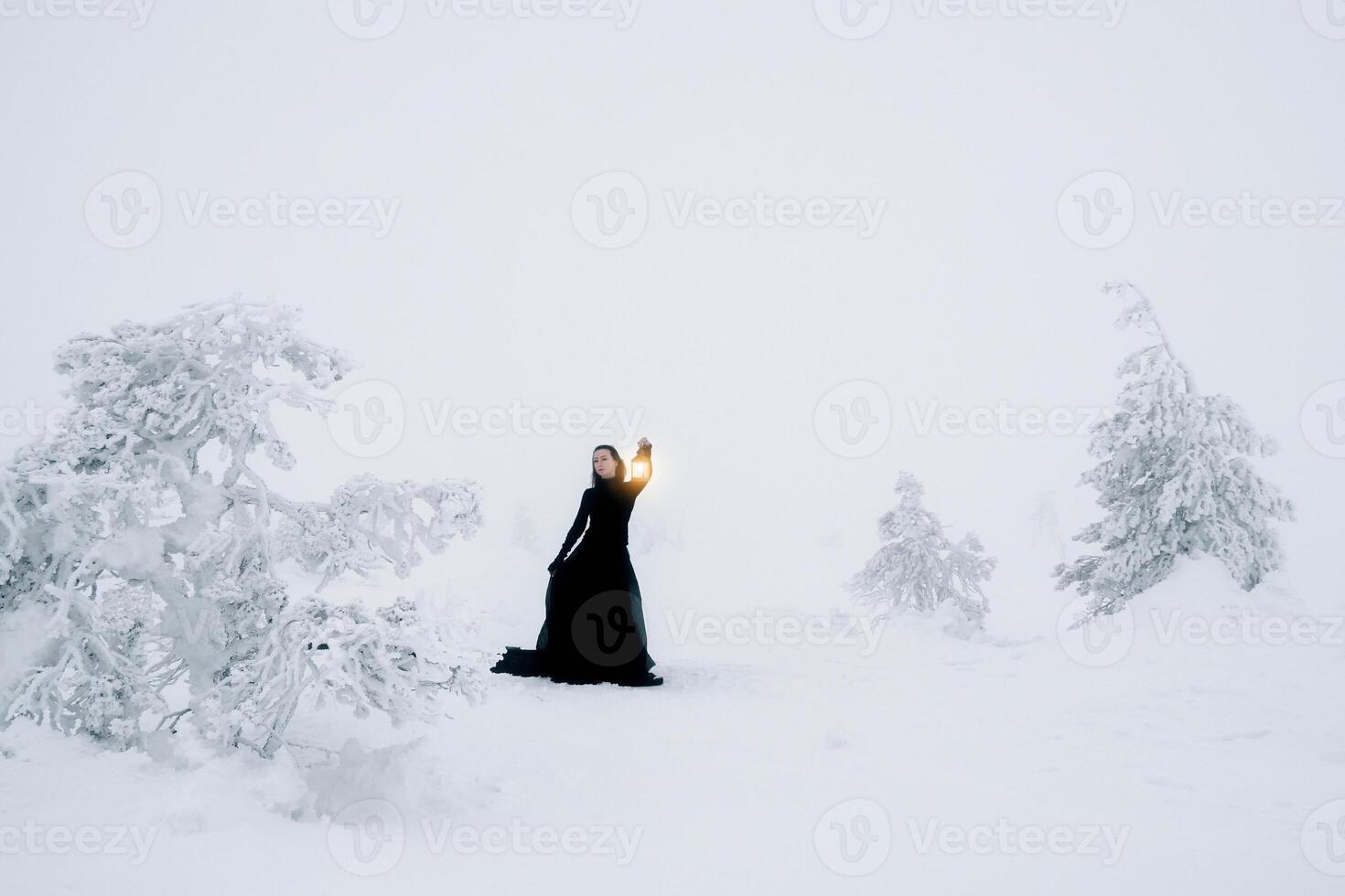 woman in long black dress with old lantern over winter hill background and snowfall. photo