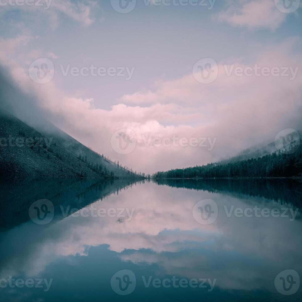 Alpine mystical natural square background with fog early morning. Silhouettes of pointy fir tops on hillside along mountain lake in dense fog. Reflection of coniferous trees in calm water. photo