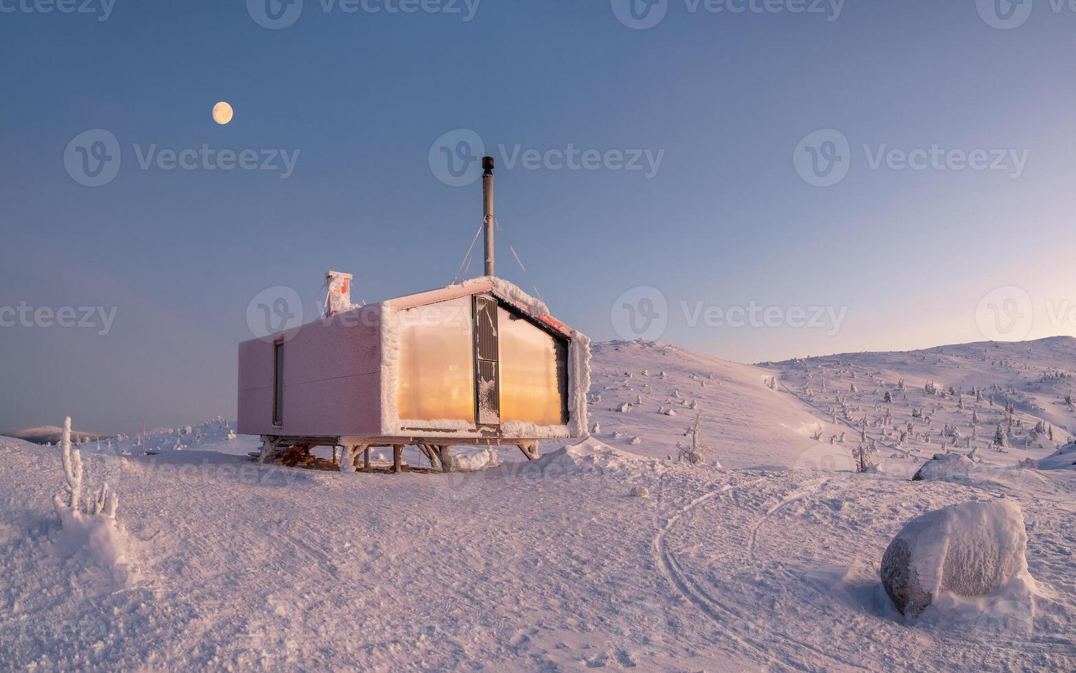 Lonely red house on a snowy frosty hill under sunrise sky with a full moon. Panoramic minimalist background with of lonely red cabin in winter. Winter holidays, comfort on a frosty night. photo