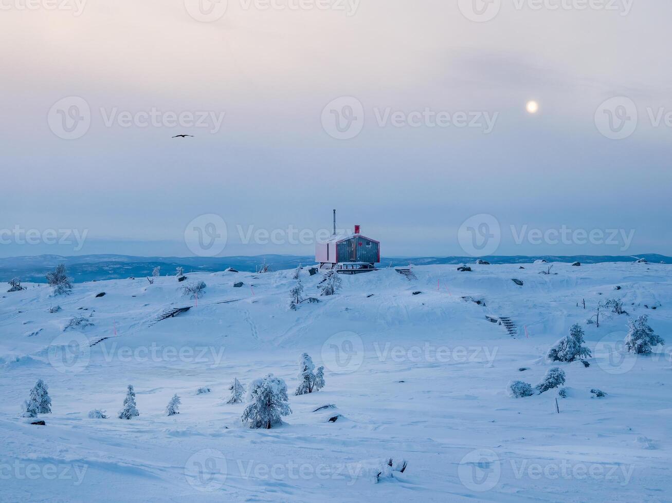 Lonely red bungalowon a snowy frosty hill under dark evening sky with a full moon. Minimalistic background with of lonely red cabin in winter. photo