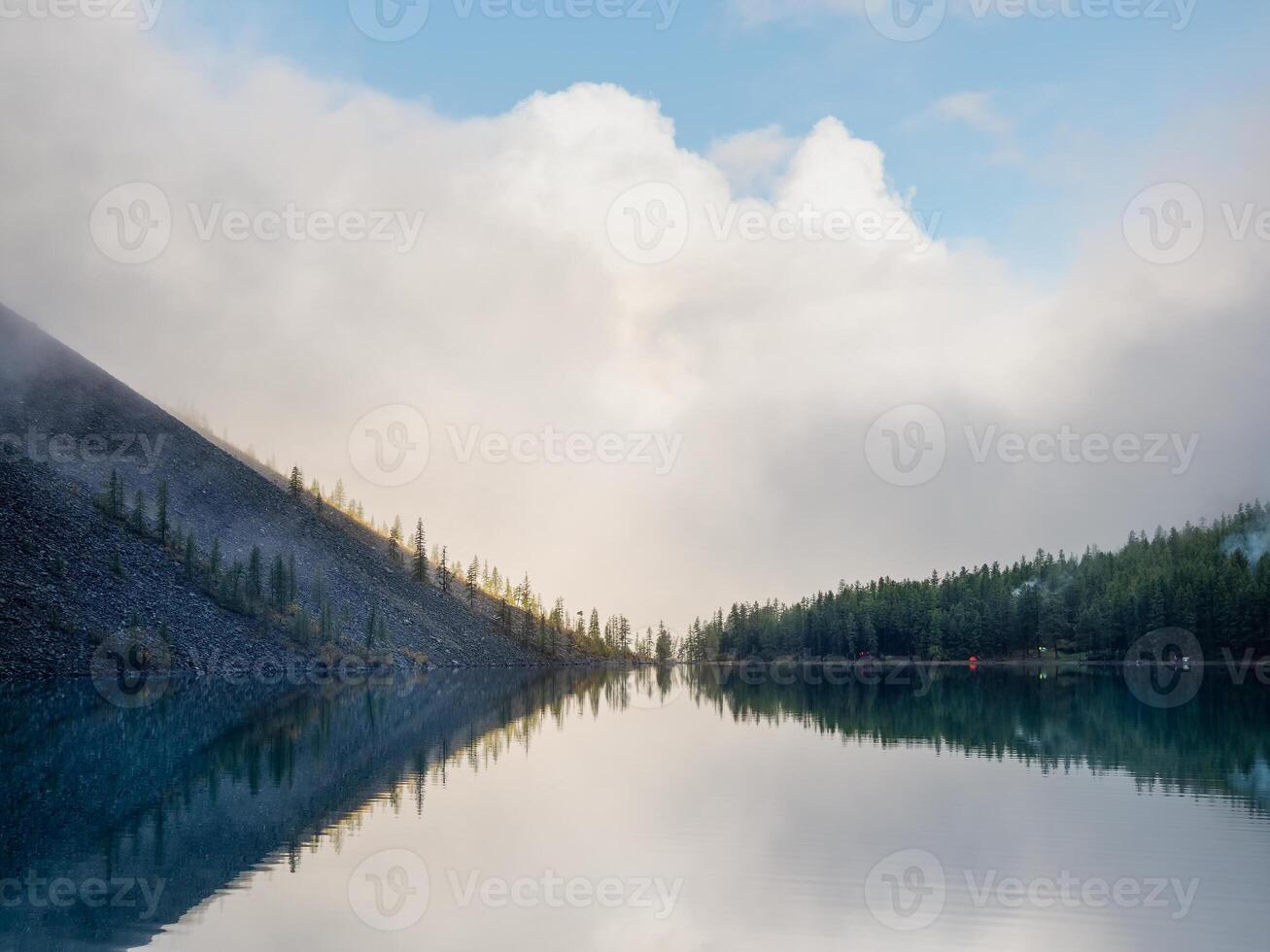 Morning silhouettes of pointy fir tops on hillside along mountain lake in dense fog. Alpine tranquil landscape at early morning with reflection in clean water. Dark ghostly atmospheric scenery. photo
