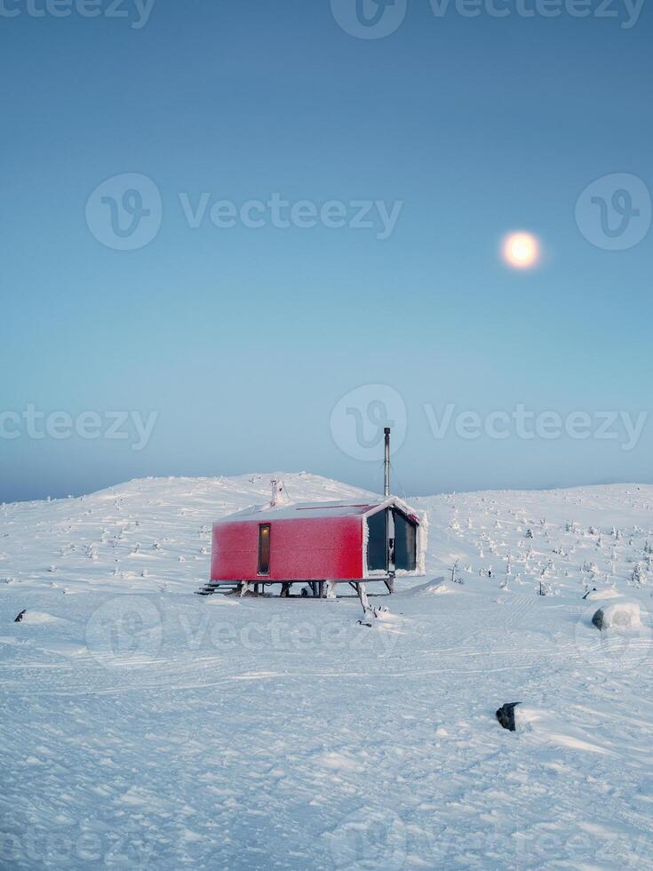 Lonely red house on a snowy frosty hill under evening sky with a full moon. Vertical minimalistic background with of lonely red cabin in winter. photo