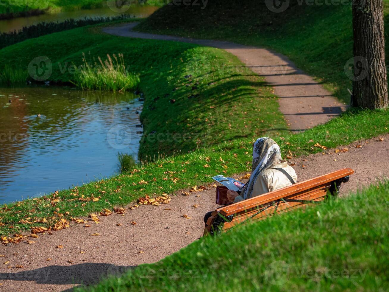 A woman in a headscarf is sitting on a bench in an autumn park and leafing through a magazine. Outdoor recreation photo