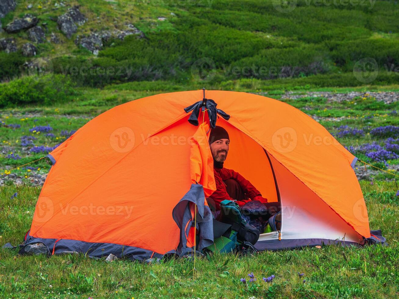 Portrait of a smiling bearded tourist sitting in a tent. Solo trekking. Camping on a summer green rocky high-altitude plateau. Peace and relaxation in nature. photo