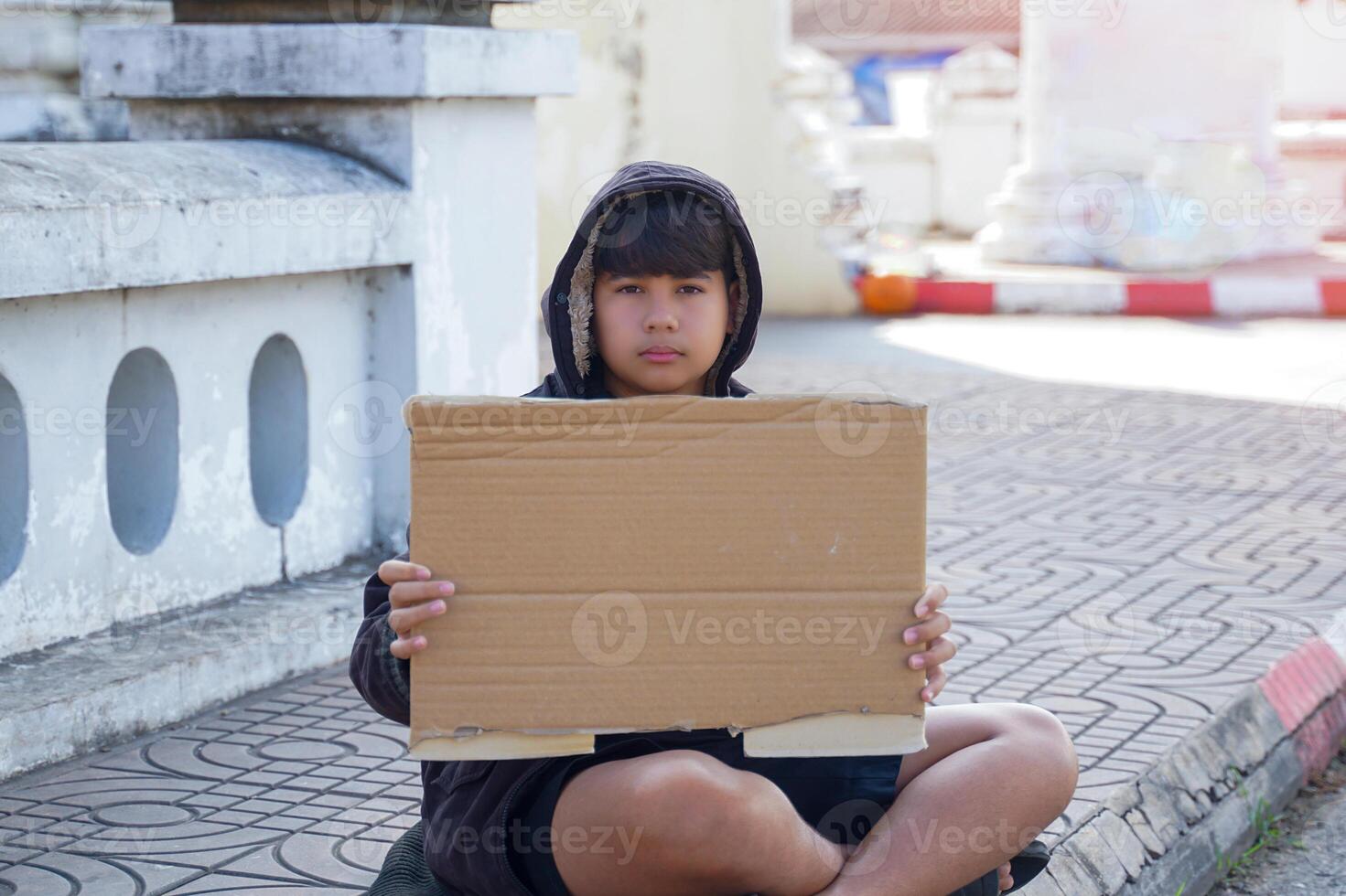 teenage Asian boy wearing a black hoodie sits alone holding a cardboard paper sign in an empty space on the sidewalk. The concept of homeless children, social problems, having problems, being lonely. photo