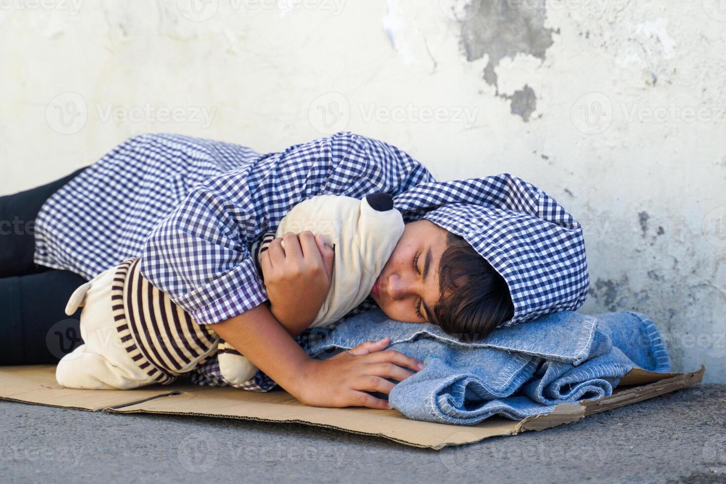 teenage boy wore a hood over his head and hugged an old doll on a piece of cardboard used to sleep against a wall on the side of the road. photo