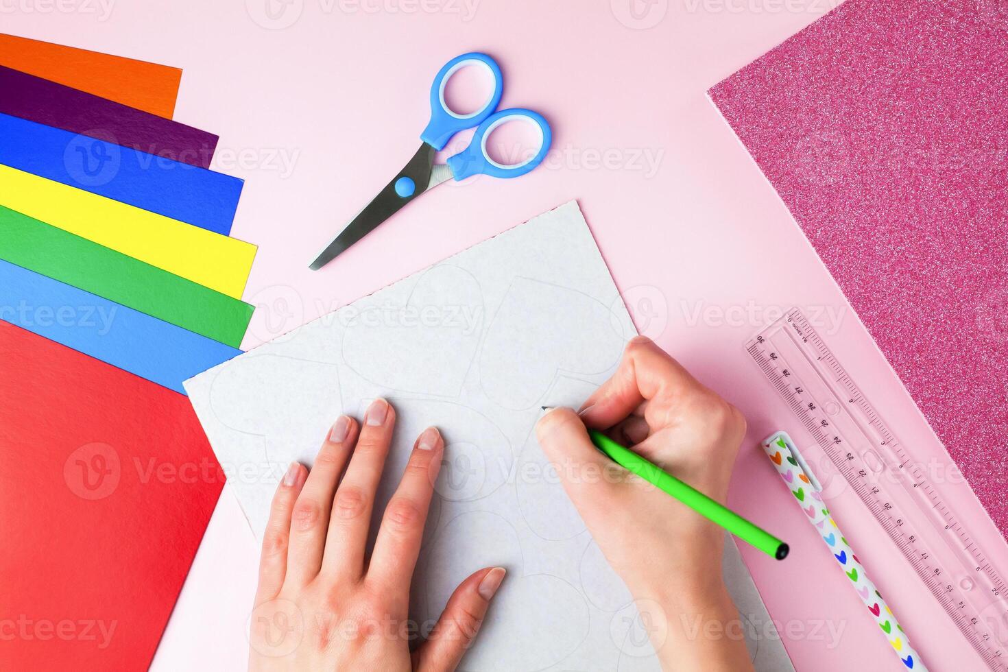 Woman's hand holds a pencil and draws a hearts on paper on pink background. DIY concept for Valentine's day card crafts. Top view. Flat lay. Step by step photo instruction. Step 2.