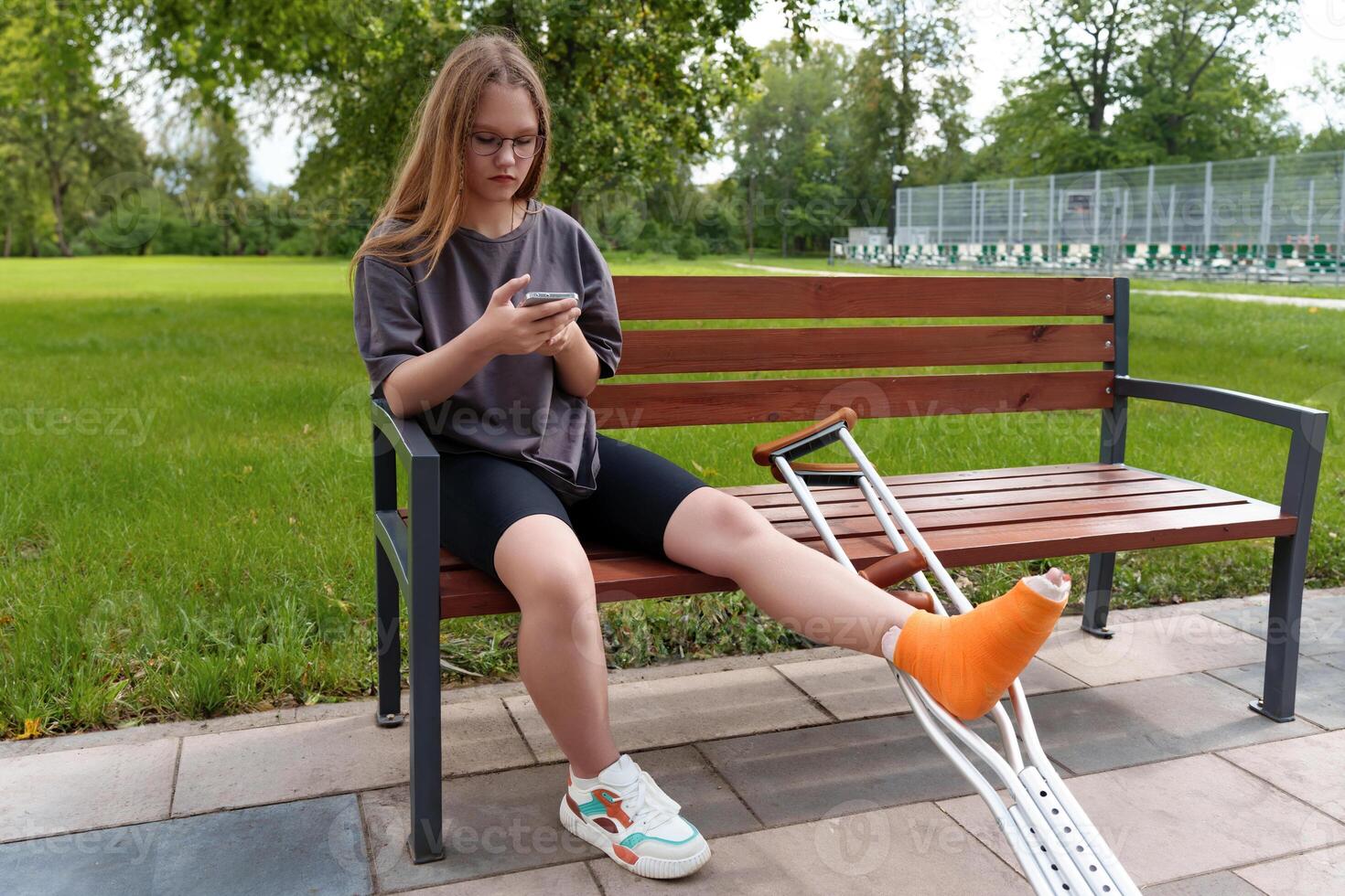 Young female with a casted leg relaxing on a bench in a lush summer park, reading content on her mobile device during her rehabilitation photo