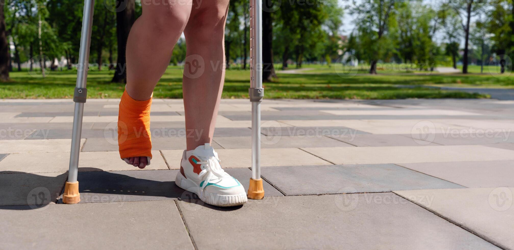 leg encased in an orange cast, with crutches visible in the lush, green background of a summer park setting. Concept of injury recovery photo