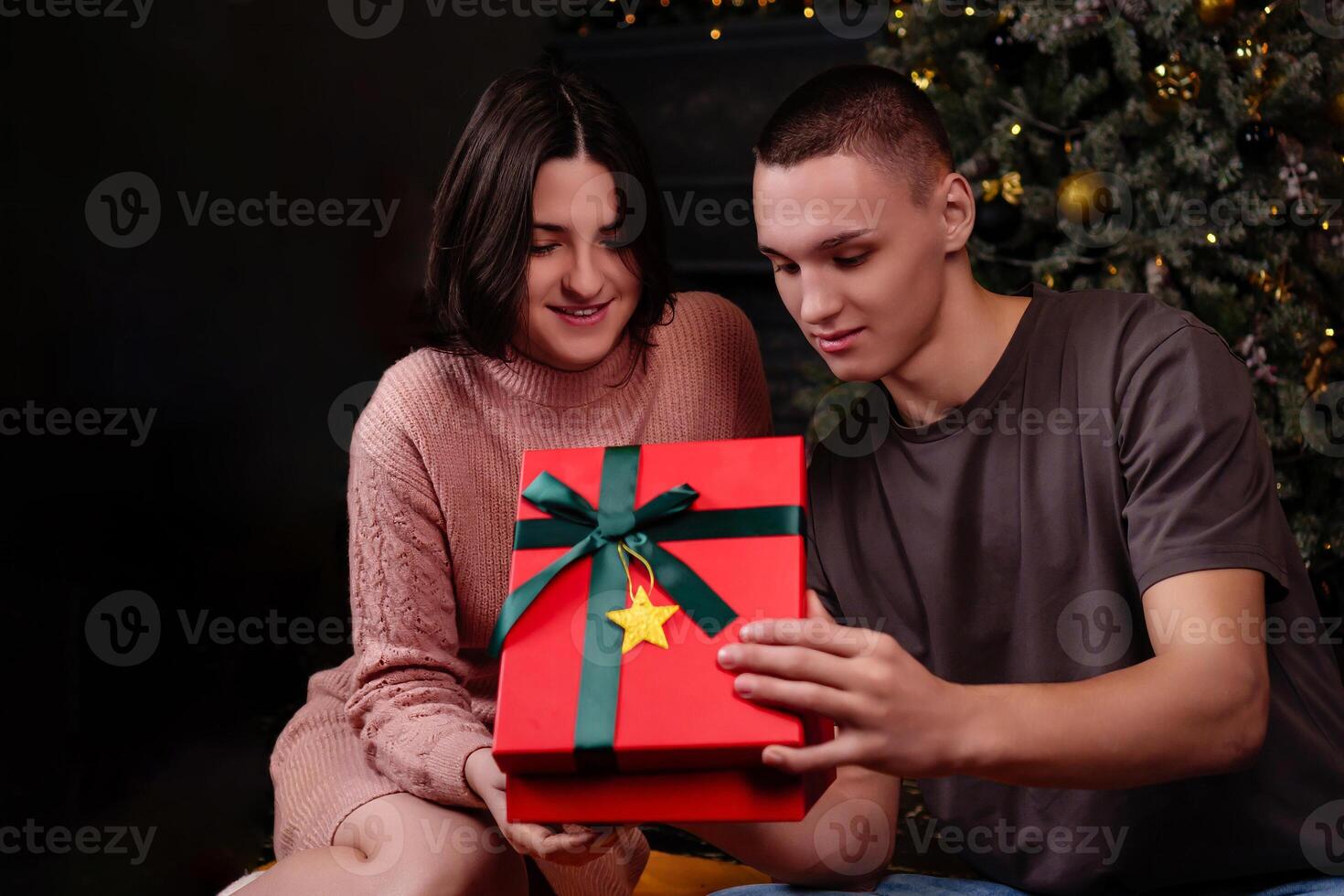 young couple in love against the background of the Christmas tree open a red gift box with interest looking inside photo