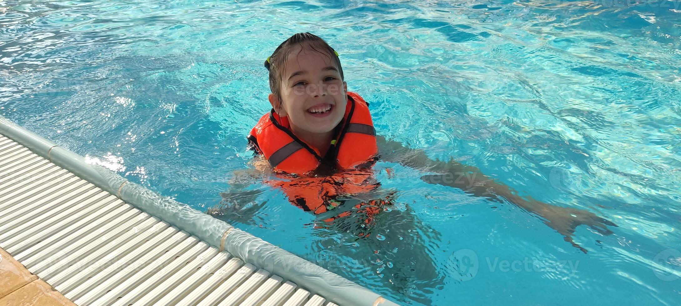 Happy child in orange life jacket enjoying a swim in clear blue pool photo