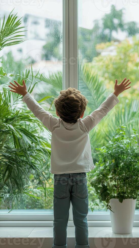 Child enjoys the beauty of nature through a large window surrounded by vibrant green plants during the peaceful morning hours photo