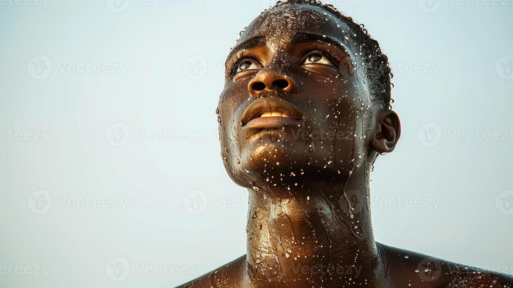 A young man stands under the sun, looking up with water droplets glistening on his skin photo