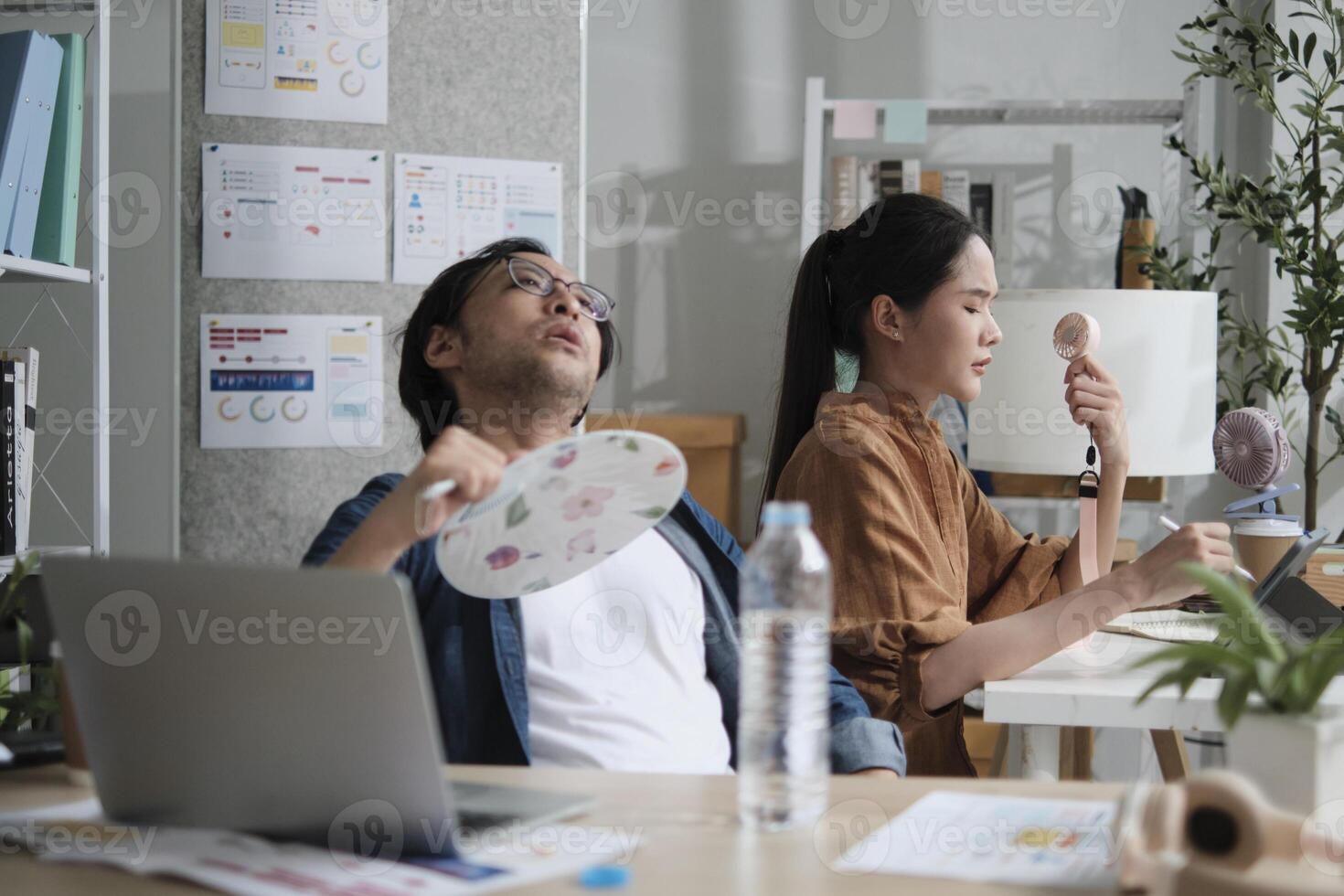 Two workers are working in hot temperatures at desk in small business office in summer, uncomfortable with no air conditioner, blowing wind with fans, drinking water, and sweating. photo