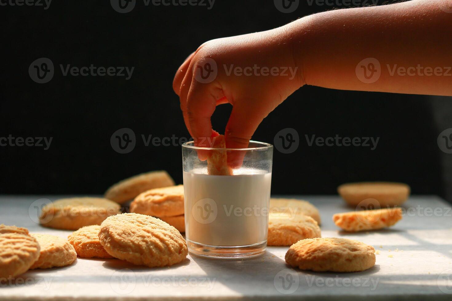 Child dipping tasty cookie in glass of milk on black background photo
