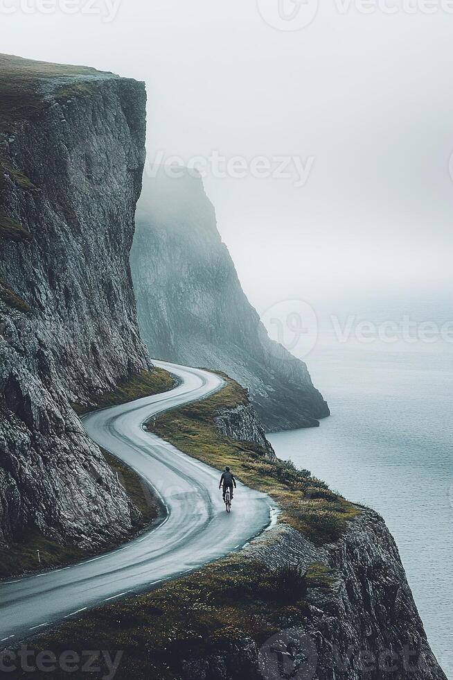 A cyclist rides along a narrow, twisting road that hugs the cliffs of a Nordic coastal landscape. The soft muted colors and mist create a tranquil atmosphere perfect for adventure seekers photo