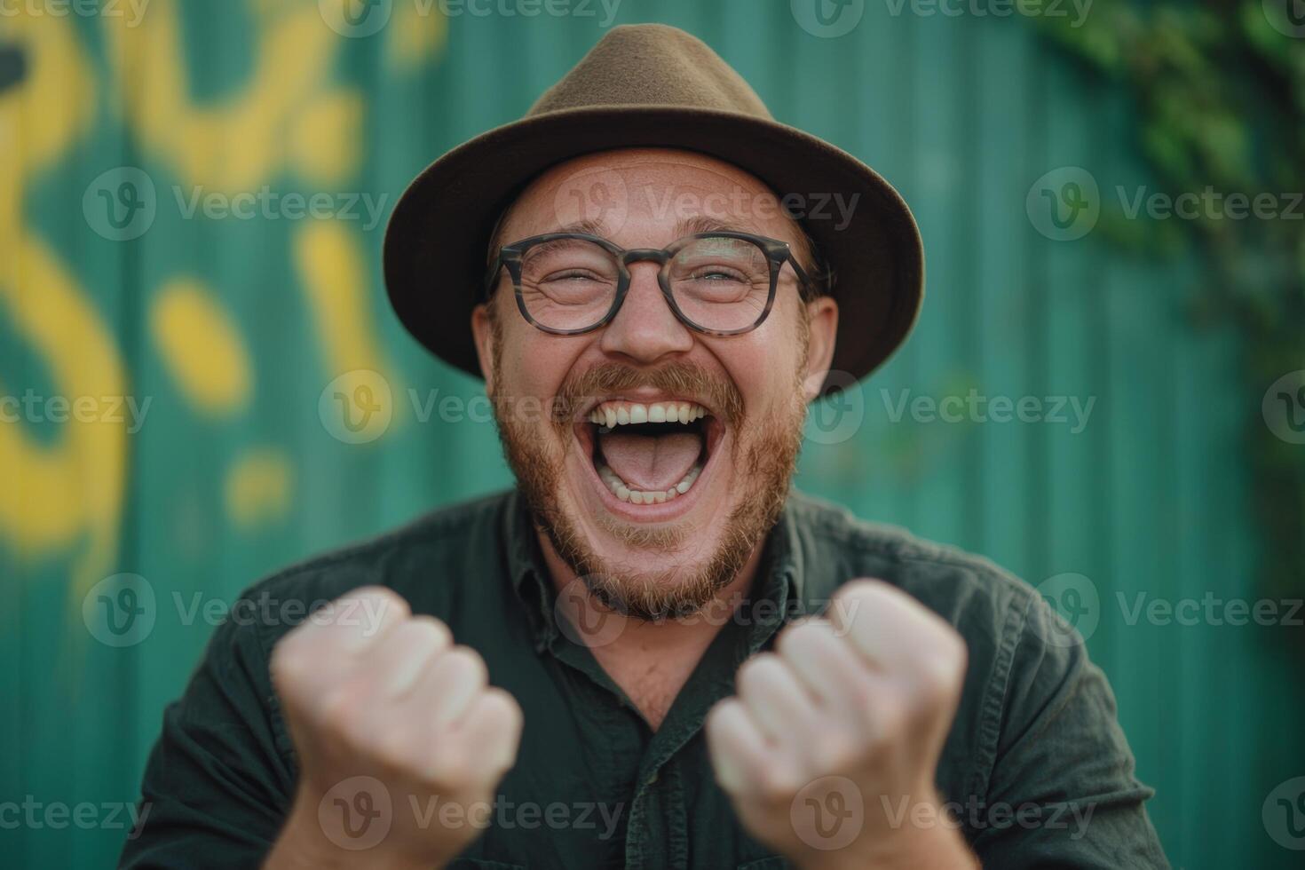 Cheerful man wearing a hat celebrates joyfully with raised hands in front of a vibrant green backdrop during daytime photo