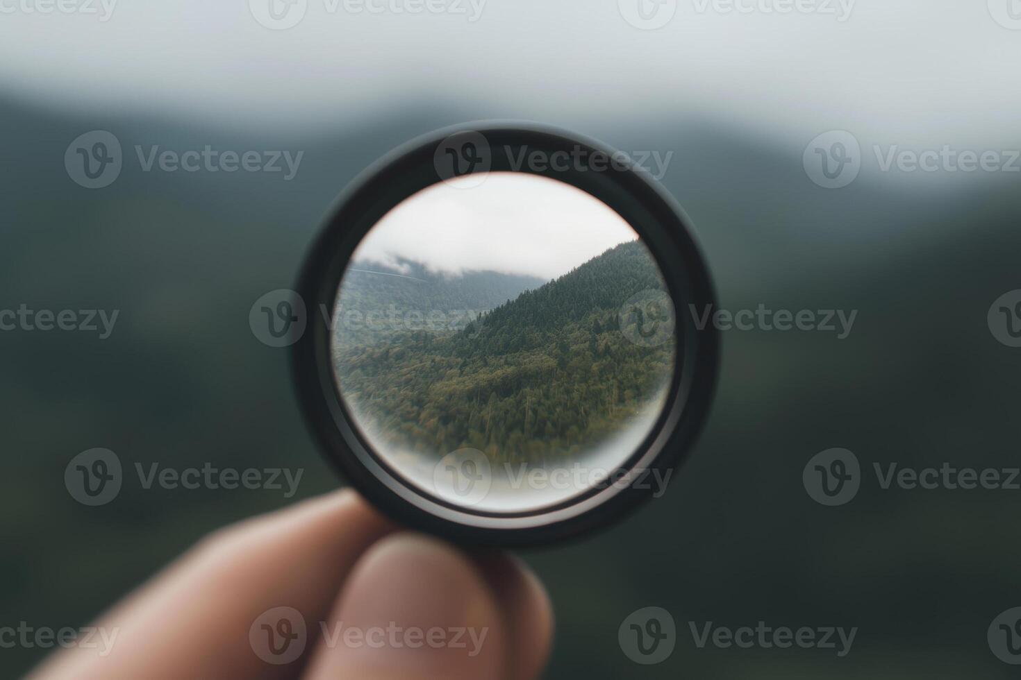 A person's hand holds a lens in front of lush green mountains under a cloudy sky in the serene countryside photo