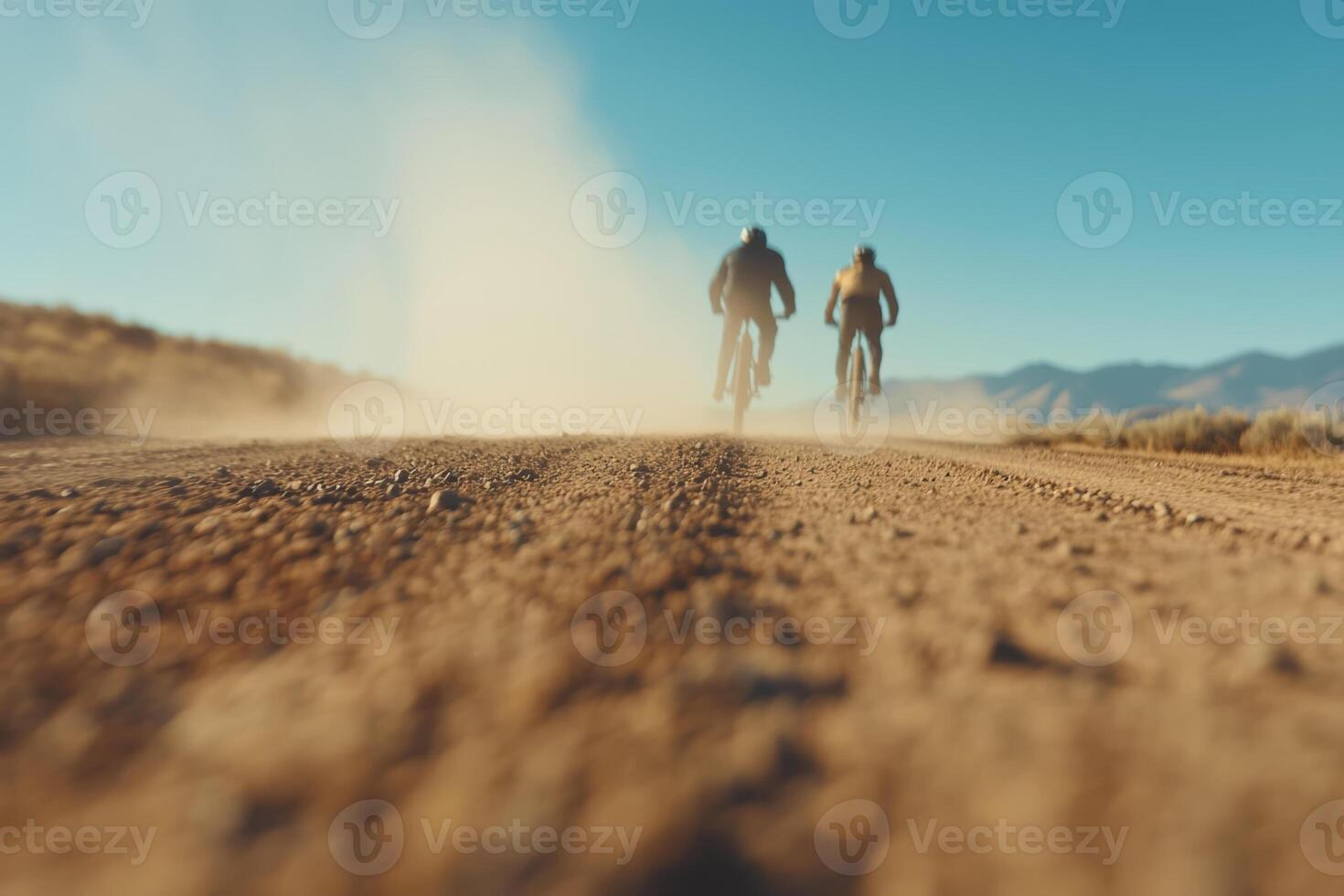 Two cyclists riding on a dusty trail surrounded by mountains in bright sunlight during a clear day photo
