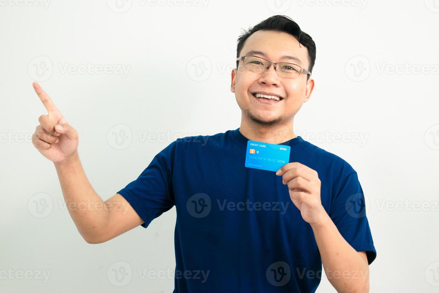 Delighted asian man blue tshirt hold credit card with pointing pose. Banking, Financial isolated background, blue color concept. photo