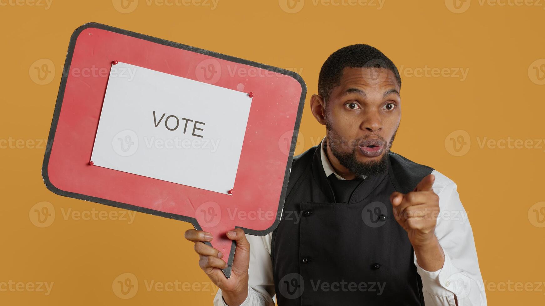 Catering employee showing a message to vote on red speech bubble, presenting the cardboard icon to do a commercial for politics. Waiter in uniform works on customer service. Camera A. photo