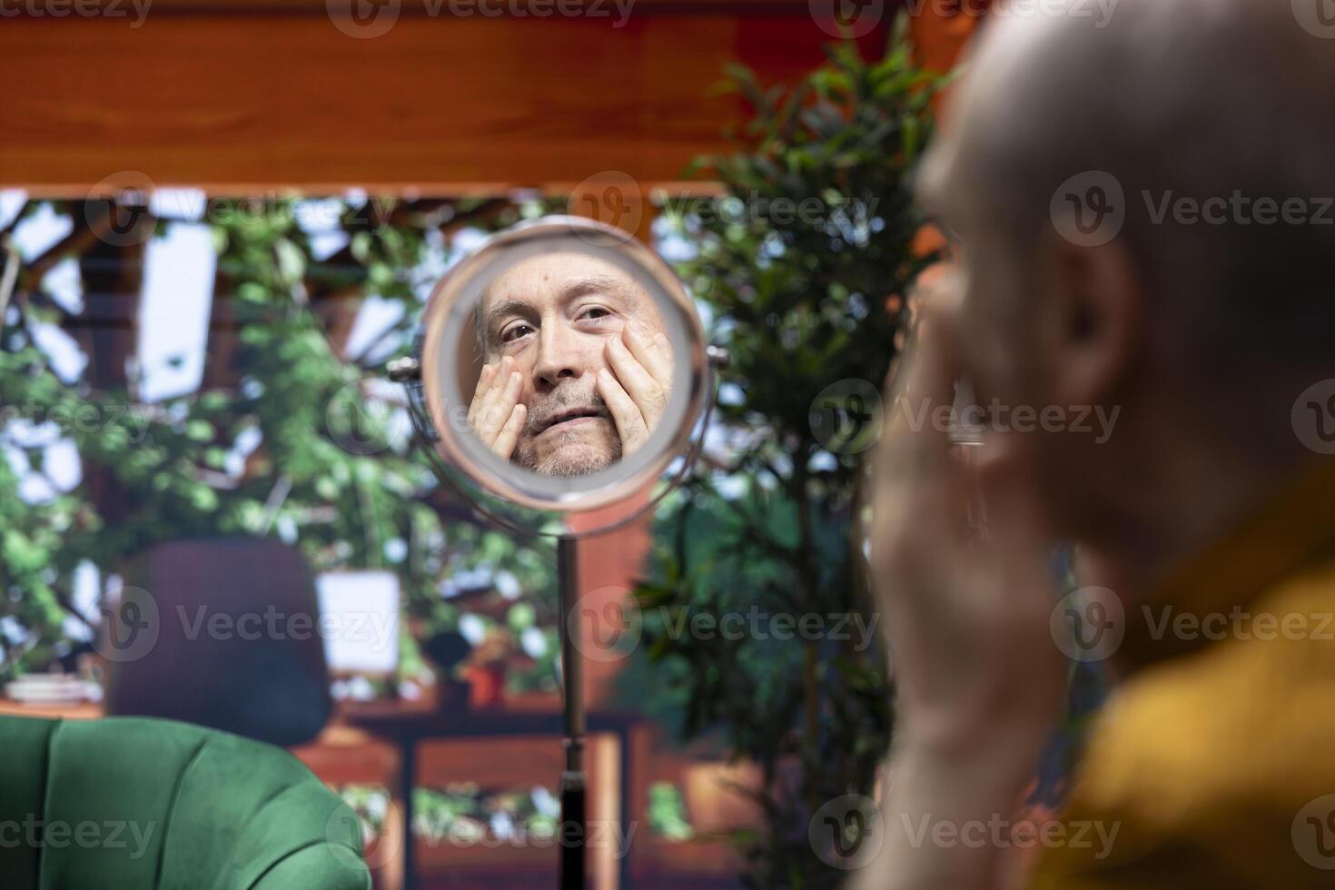 Pensioner using serum and cream to preserve his mature skin with wrinkles, looking in the mirror at his aging face. Doing a pampering daily routine for personal care, grooming products. photo