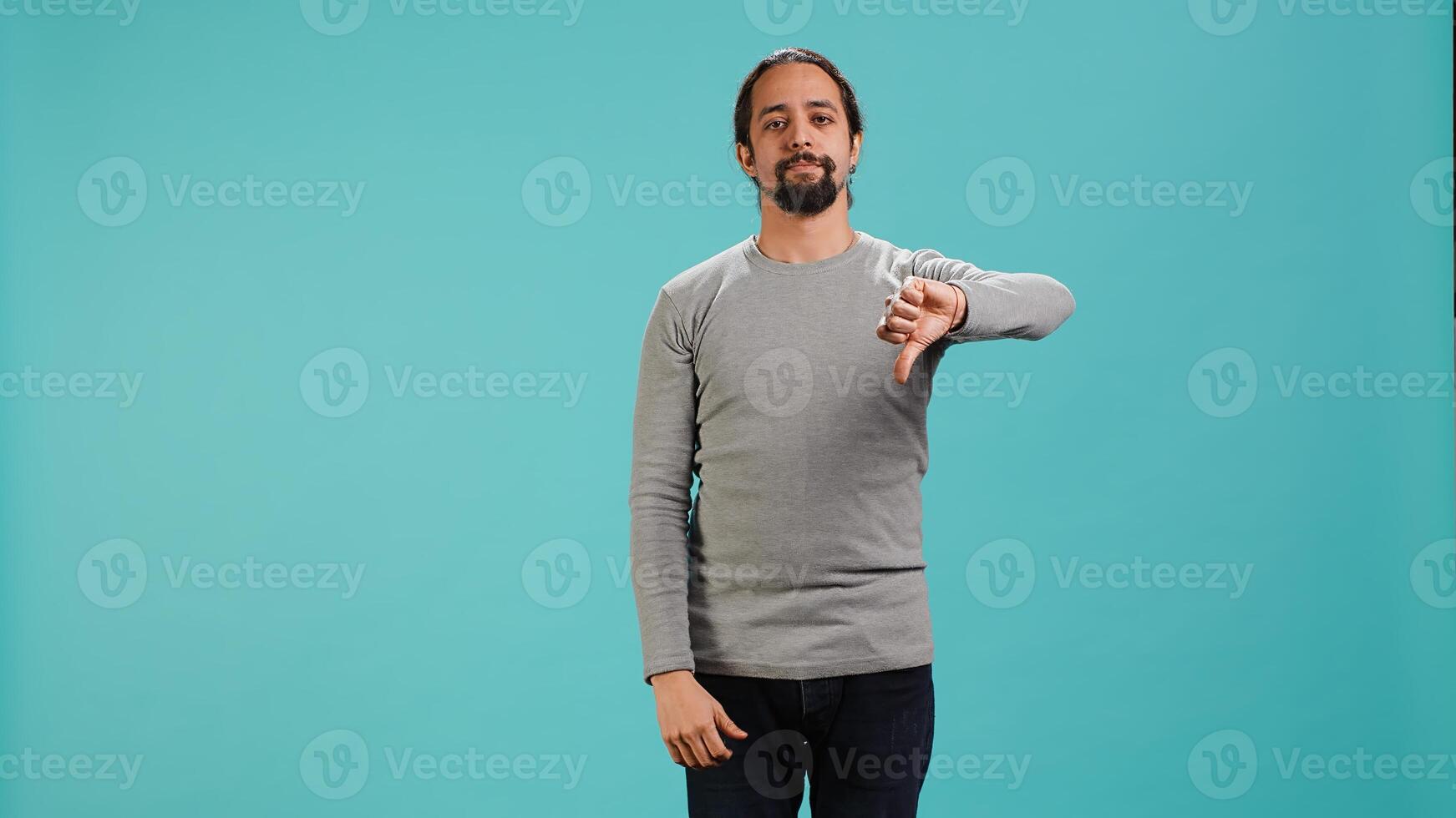 Upset man showing thumbs down sign gesturing, being disapproving. Disappointed Arab person against gaming, doing rejection hand gesture, isolated over studio background, camera A photo