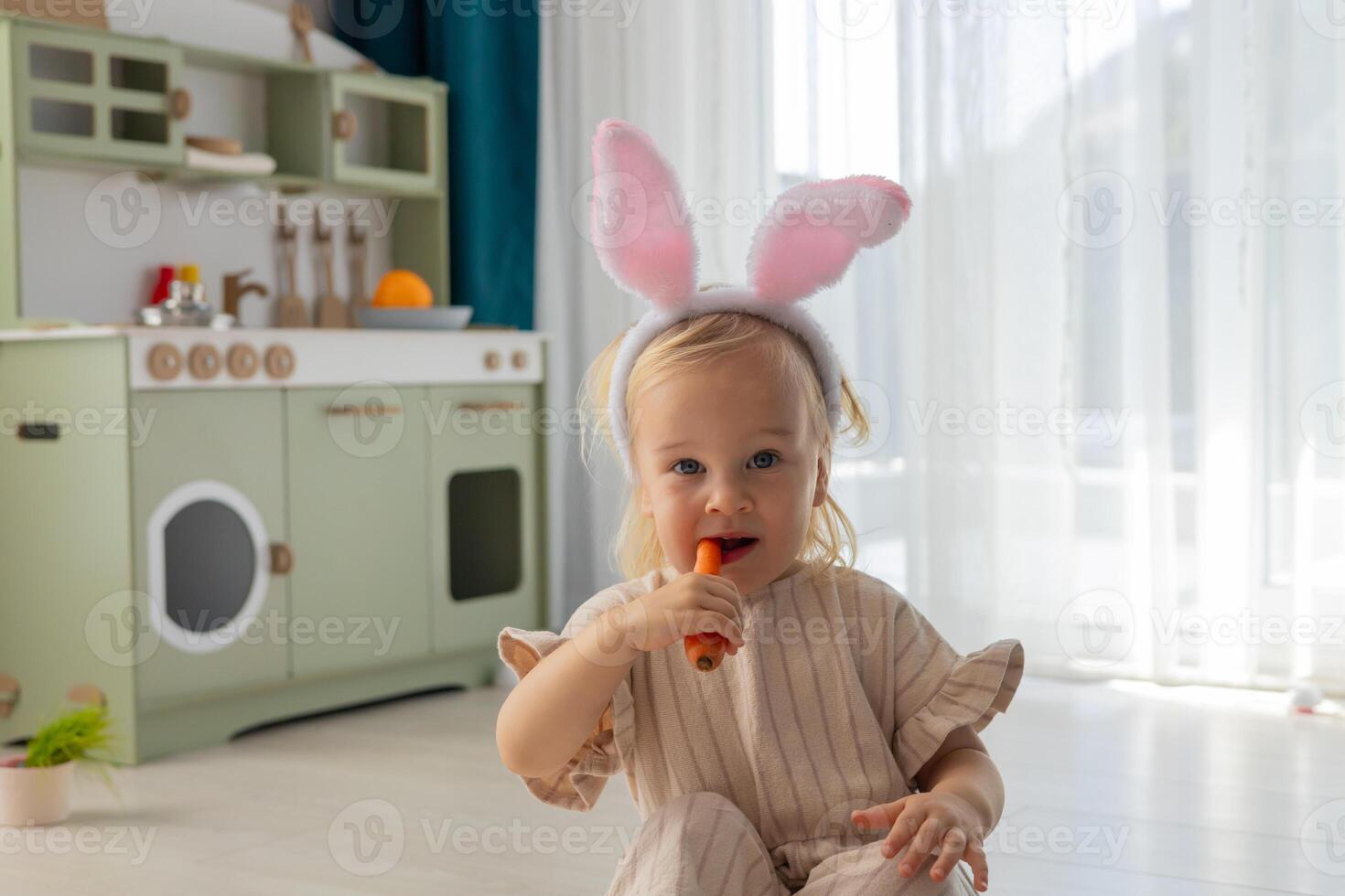 Portrait of a happy toddler girl in bunny ears eating a carrot. photo
