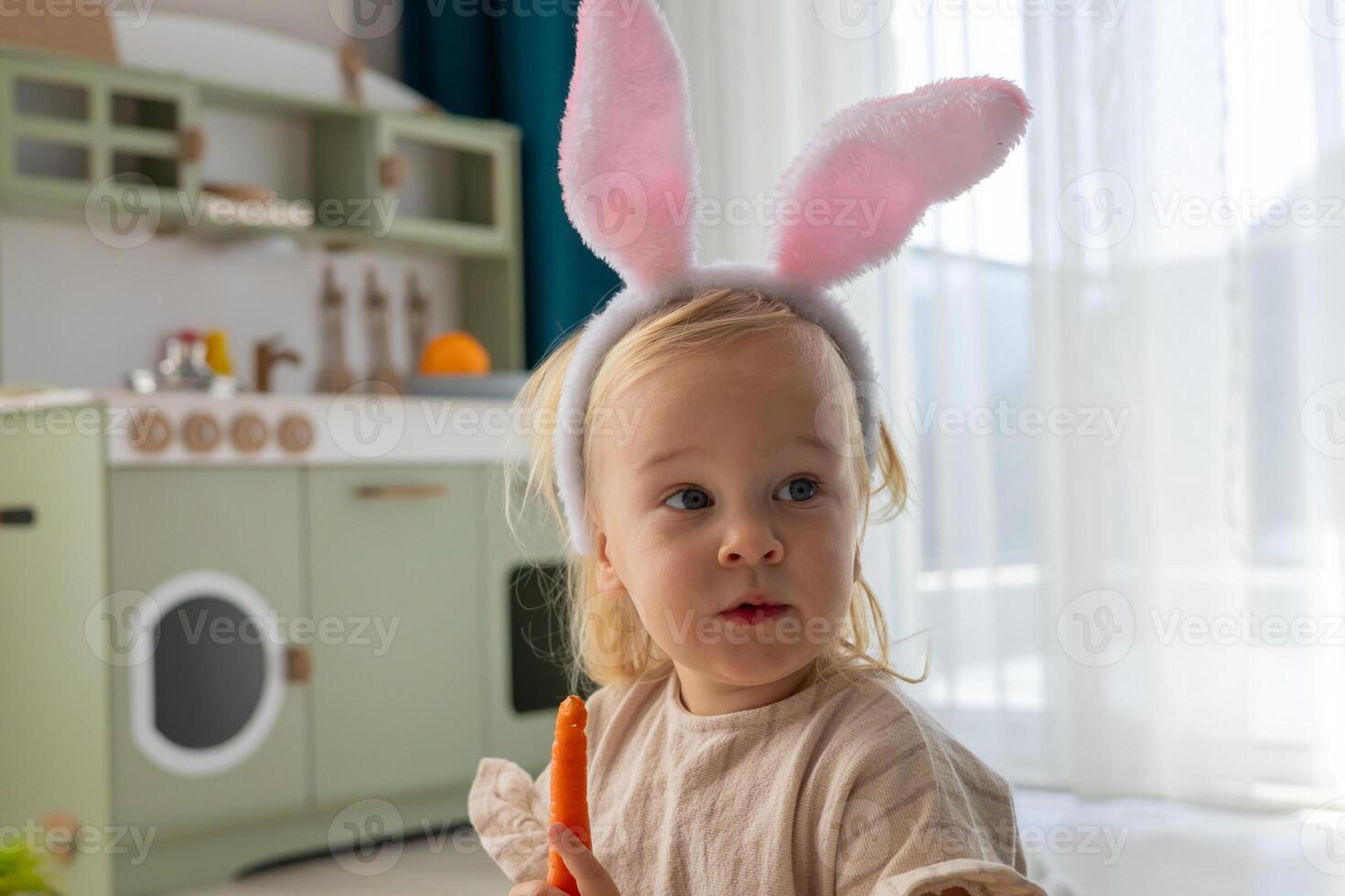 Portrait of a happy toddler girl in bunny ears eating a carrot. photo