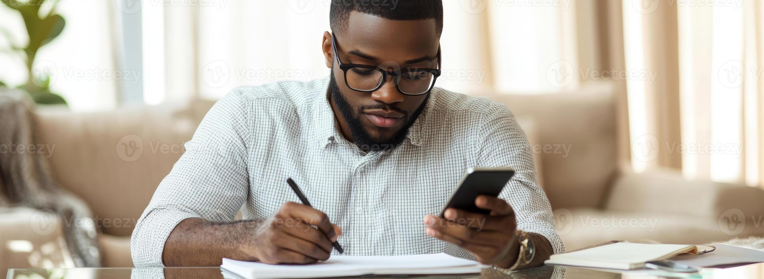 A man with glasses and a beard is writing on a notepad while looking at his phone photo