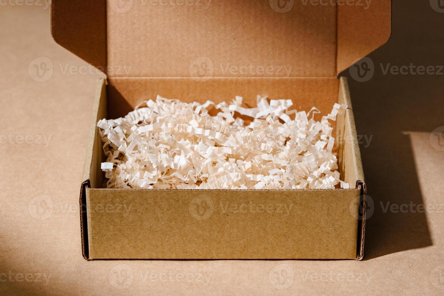 Open cardboard box with shredded paper packaging resting on a light brown surface illuminated by soft natural light photo