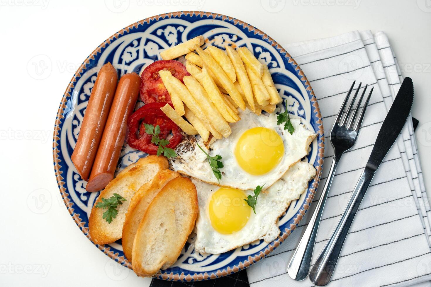 Breakfast plate with fried eggs, sausage, tomatoes, and fries served on a decorative blue plate photo