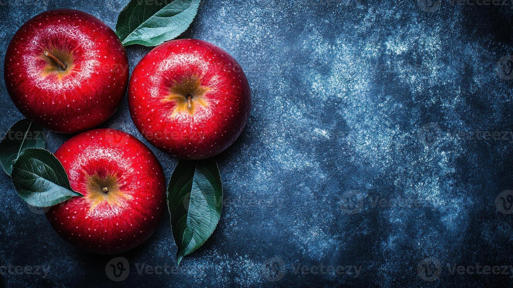 Three shiny red apples with green leaves are artistically placed on a dark blue backdrop photo