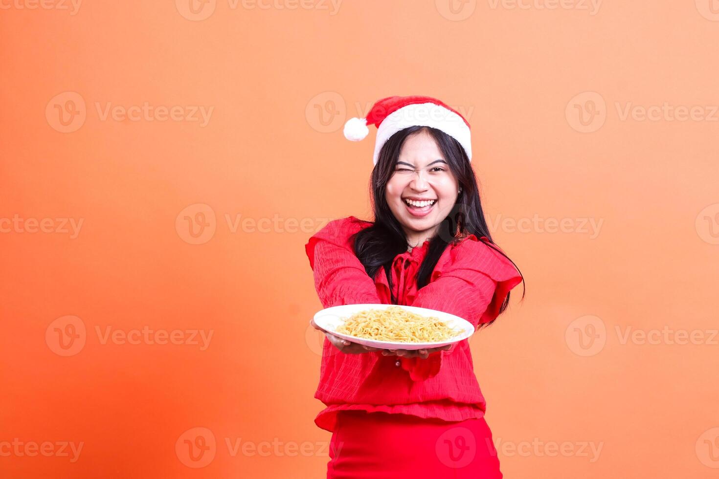 expression of teenage girl wearing christmas dress, santa hat, cheerful winking looking at camera, both hands holding white plate filled with noodles in front while offering, isolated on background photo