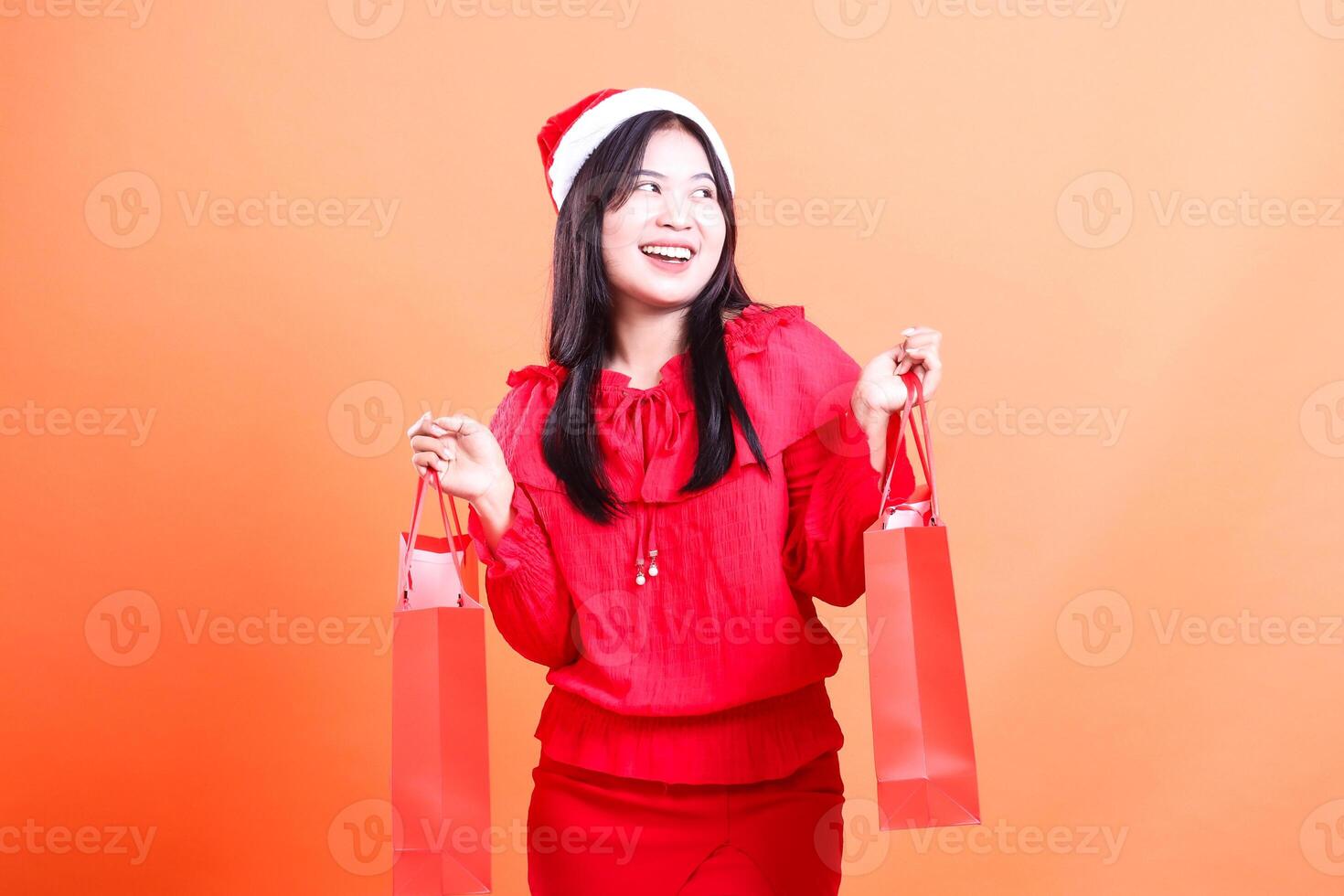 Asian young woman expression wearing red sweater and santa hat, standing cheerful to the left both hands lifting red bag of christmas gifts, isolated on orange background photo